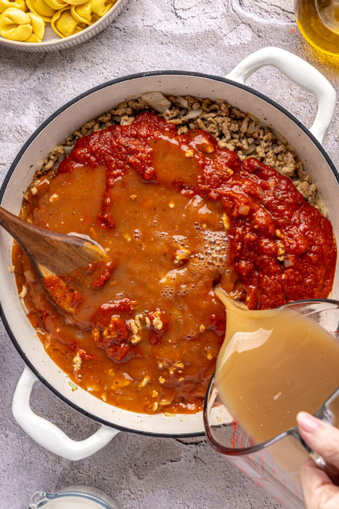 broth and marinara sauce being added to a pot of cooked sausage and onion.