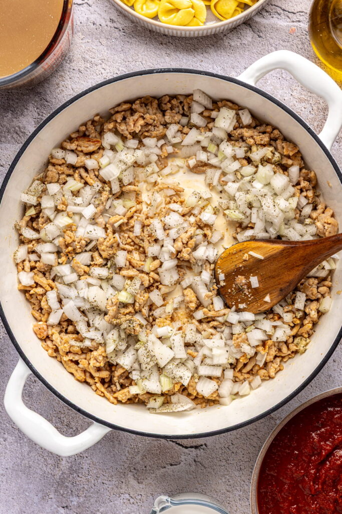 onion and seasonings being stirred into cooked sausage in a pot.