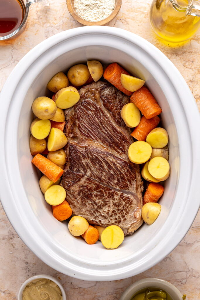seared chuck roast in a slow cooker surrounded by raw carrots and potatoes